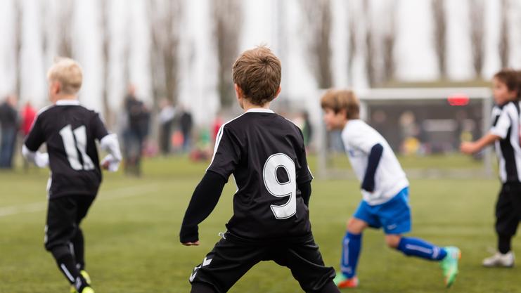 Kinder spielen Fußball auf einem Sportplatz; ein Spieler trägt ein schwarzes Trikot mit der Nummer 9.