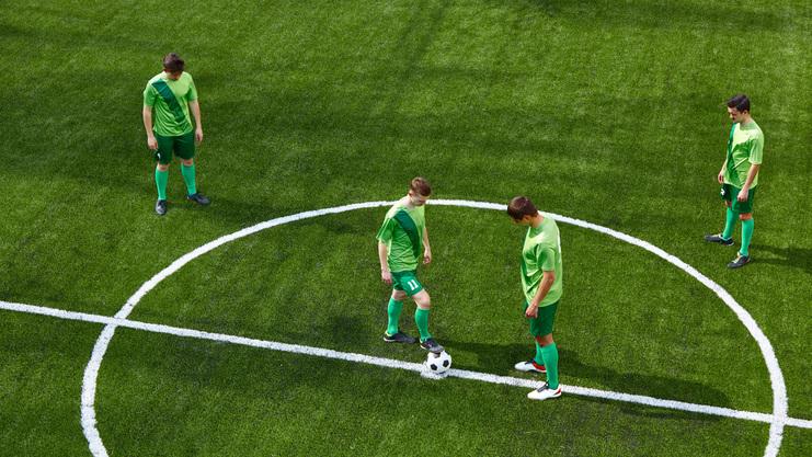 Vier Fußballspieler in grünen Trikots stehen auf einem Kunstrasenfeld, während ein Ball in der Mitte wartet.